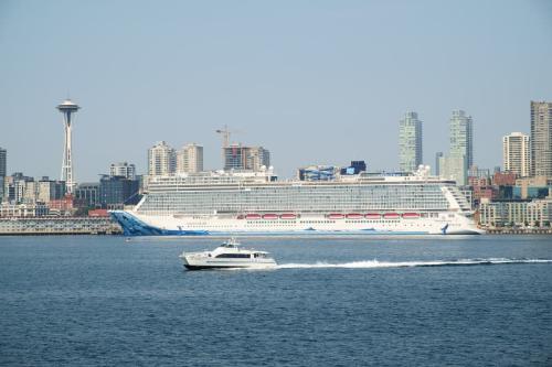 A large cruise ship passes the Seattle waterfront and Space Needle as a small passenger ferry crosses in front.