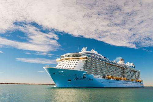 Royal Caribbean's Ovation of the Seas cruise ship sails under a partly cloudy sky with its bright yellow lifeboats visible.