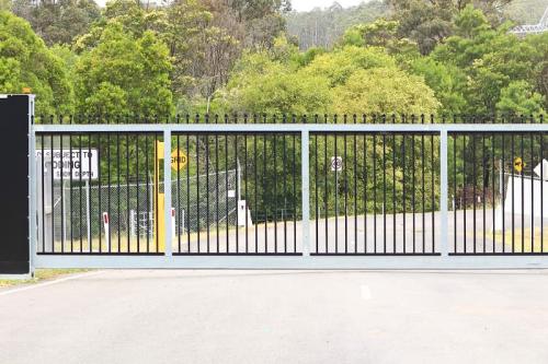 A secured black metal automated gate with fencing controls entry to a paved lot surrounded by trees.